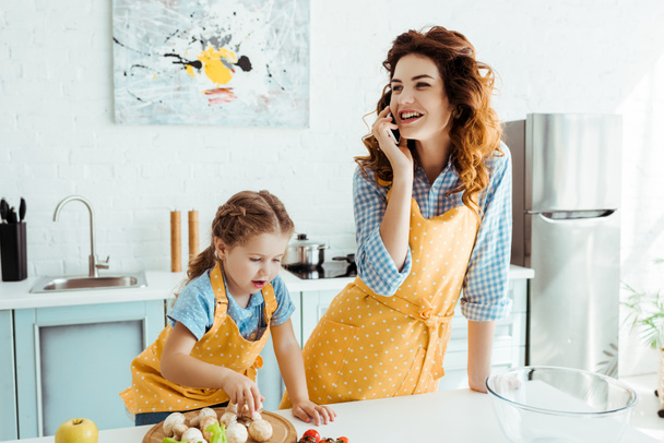 happy mother talking on smartphone while daughter putting mushrooms on chopping board - Fotoğraf, Görsel