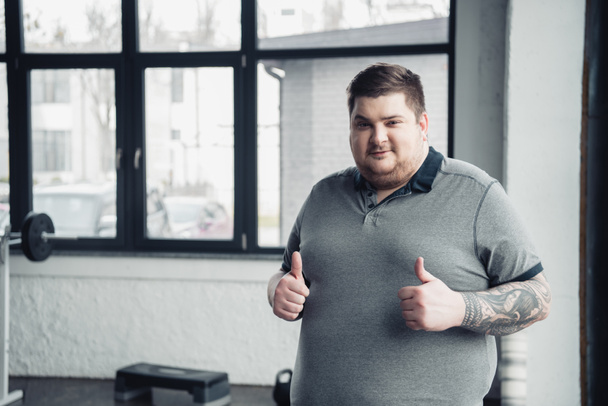 Человек с избыточным весом с татуировкой смотрит в камеру и показывает большие пальцы в спортзале с копировальным пространством
 - Фото, изображение