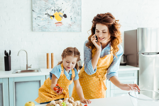 happy mother talking on smartphone while daughter holding cherry tomatoes - 写真・画像