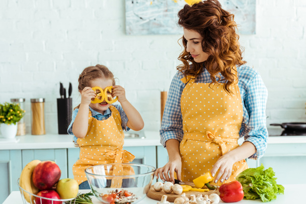 Mutter in gepunkteter gelber Schürze gibt Blick auf Tochter mit Paprika-Scheiben in Augennähe - Foto, Bild