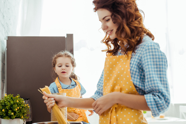 curious daughter looking at mother in polka dot yellow apron putting raw spaghetti in pot - Photo, image
