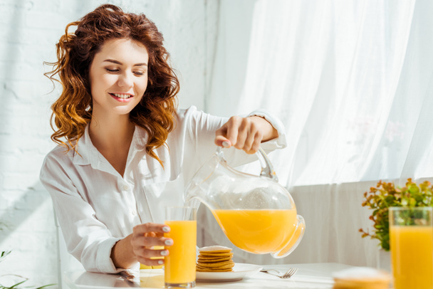 beautiful happy woman sitting at table and pouring orange juice in glass while having breakfast - Fotografie, Obrázek