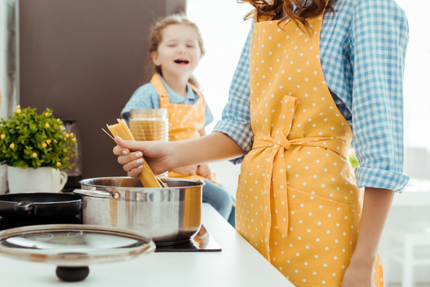 cropped view of woman in polka dot yellow apron putting raw spaghetti in pot with excited daughter on background - Photo, Image
