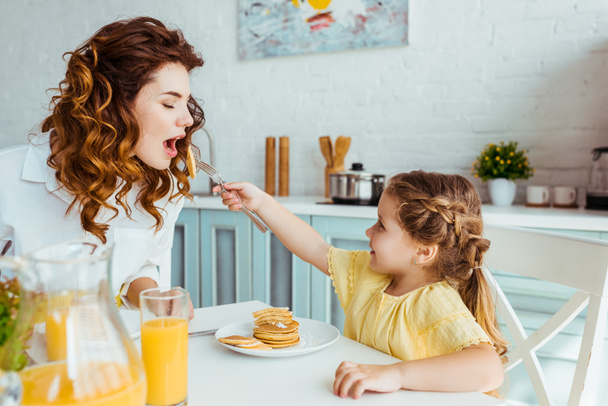 smiling daughter feeding mother with pancakes during breakfast - Foto, Imagem