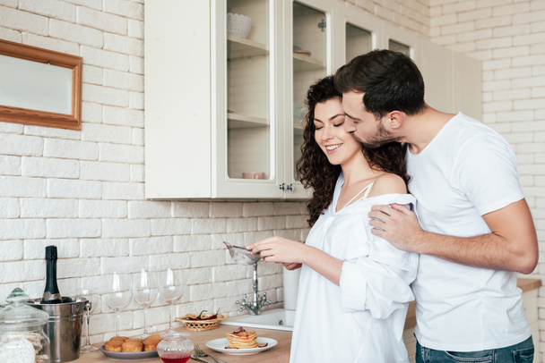 man kissing girlfriend while she preparing breakfast in kitchen - Foto, Bild