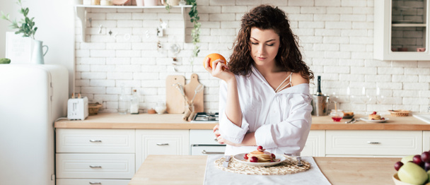 panoramic shot of girl holding orange and looking at pancakes in kitchen  - Fotoğraf, Görsel