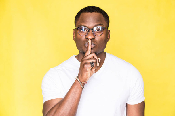 Portrait of a young handsome man in glasses showing a gesture of silence with a finger to his lips on a yellow background. - Fotografie, Obrázek