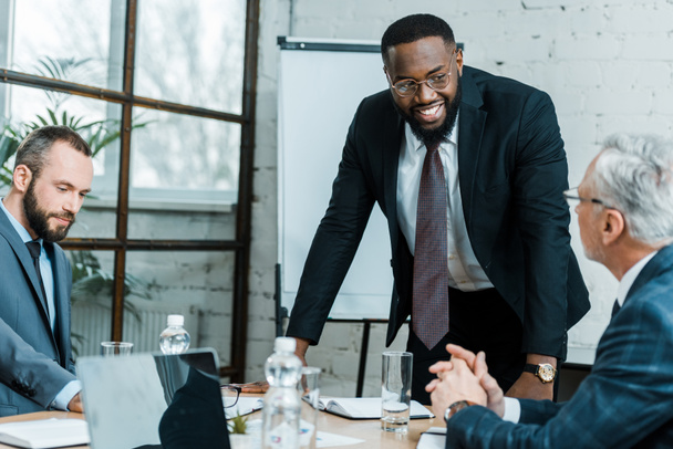 foco seletivo de alegre treinador de negócios afro-americano sorrindo perto de colegas de trabalho
  - Foto, Imagem