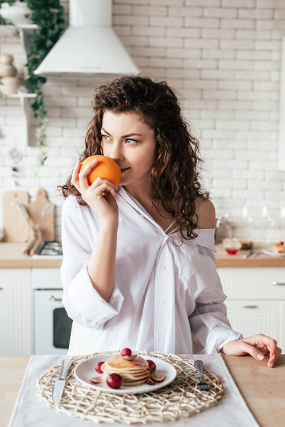 sexy girl in white shirt holding orange and looking away in kitchen - Фото, изображение