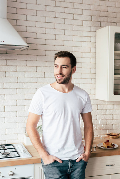 smiling man in jeans standing with hands in pockets and looking away in kitchen - 写真・画像