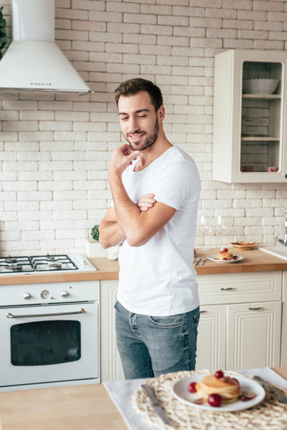 bearded man in jeans looking at pancakes with smile in kitchen - Фото, изображение