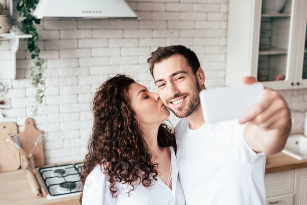 curly girl kissing boyfriend while he taking selfie in kitchen - Photo, Image