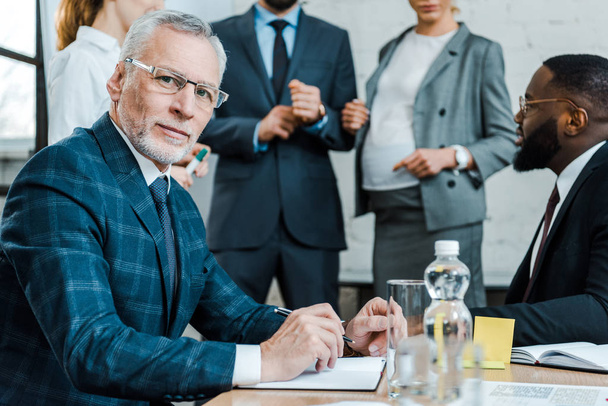selective focus of bearded businessman in eye glasses looking at camera near multicultural coworkers  - Foto, Bild