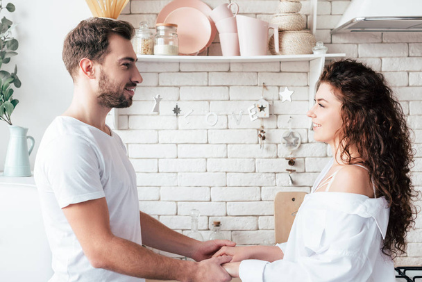 smiling couple holding hands and looking at each other in kitchen - Фото, изображение
