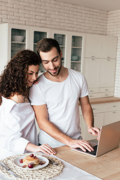 smiling couple using laptop during breakfast in kitchen - Photo, Image