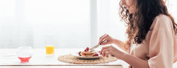 Panoramaaufnahme einer lockigen Frau, die in der Küche Pfannkuchen isst - Foto, Bild