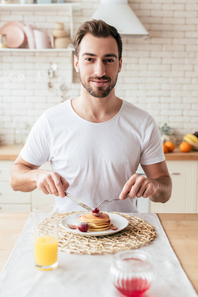 smiling bearded man in white t-shirt eating pancakes in kitchen - Fotografie, Obrázek