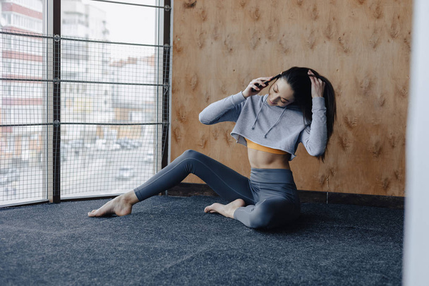 Menina fitness atraente jovem sentado no chão perto da janela no fundo de uma parede de madeira, descansando em aulas de ioga e falando ao telefone
 - Foto, Imagem