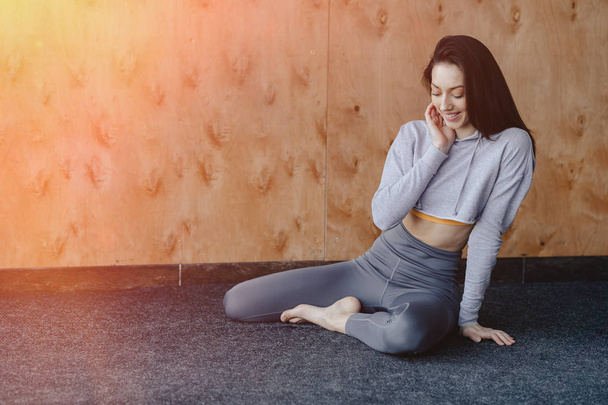 Молодая привлекательная фитнес-девушка сидит на полу возле окна на фоне деревянной стены, отдыхает на занятиях йогой
 - Фото, изображение