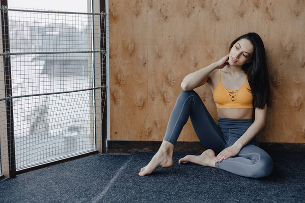 Young attractive fitness girl sitting on the floor near the window on the background of a wooden wall, resting on yoga classes - Foto, Imagem