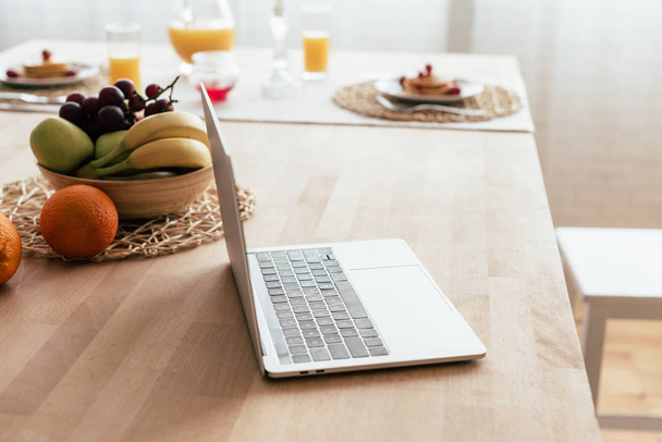 laptop en kom met vers fruit op tafel in de keuken - Foto, afbeelding