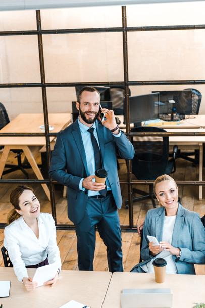 overhead view of happy bearded businessman talking on smartphone near attractive coworkers  - Photo, Image