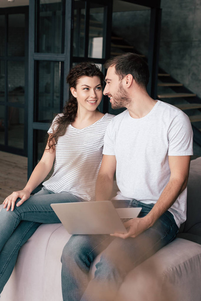 smiling couple with laptop sitting on sofa and looking at each other - Photo, image