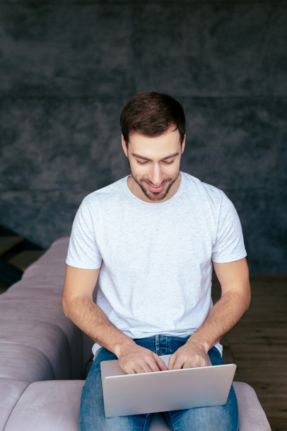 smiling bearded man in jeans sitting on sofa and using laptop - Foto, Bild