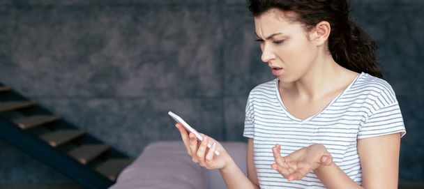 panoramic shot of irritated woman holding smartphone at home - Photo, image