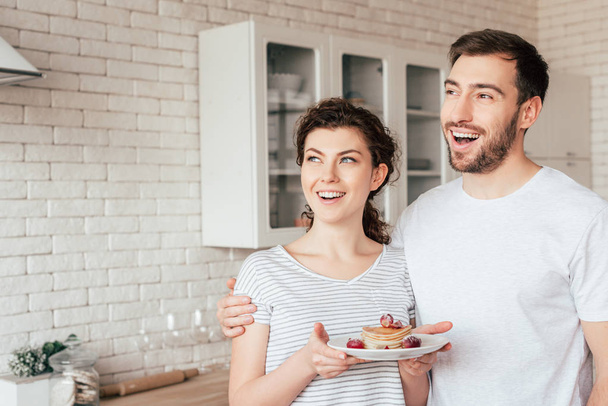 smiling man embracing girlfriend and looking away in kitchen - Foto, afbeelding