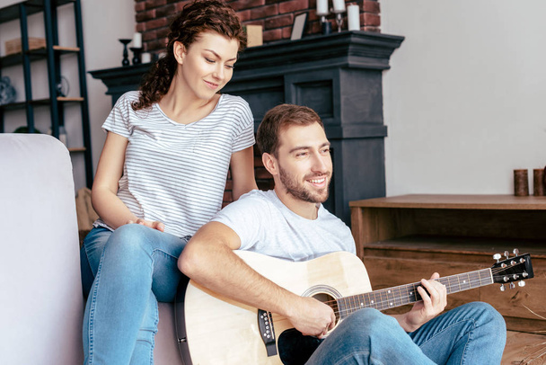 smiling bearded man playing acoustic guitar to girlfriend in living room - Fotó, kép