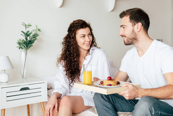 smiling couple with tray with breakfast looking at each other in bedroom - Foto, Imagem