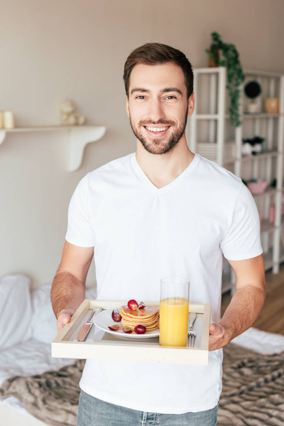 smiling man holding tray with pancakes and glass of orange juice in bedroom - Zdjęcie, obraz