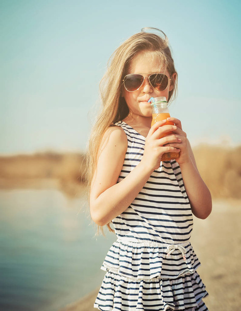 Little cute girl walking on the beach and drinking orange juice. - Photo, Image