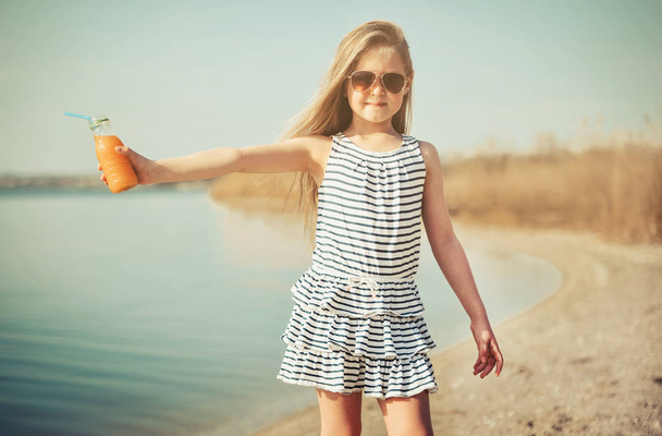 Little cute girl walking on the beach and drinking orange juice. - Photo, Image