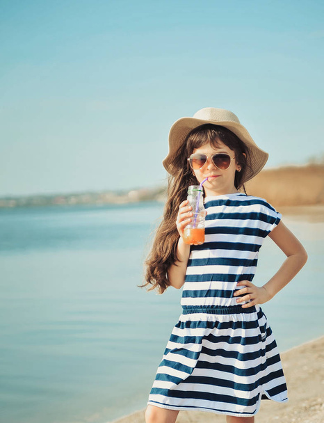 Το κοριτσάκι με το καπέλο περπατάει στην παραλία και πίνει χυμό πορτοκαλιού. - Φωτογραφία, εικόνα