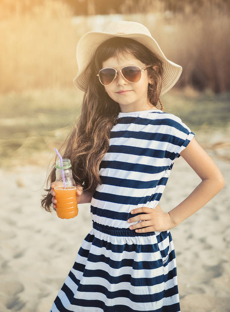 Niña con sombrero caminando por la playa y bebiendo jugo de naranja
. - Foto, Imagen