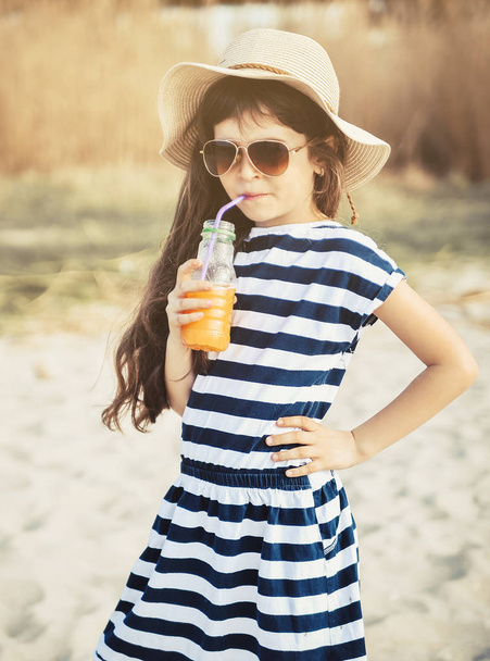 Το κοριτσάκι με το καπέλο περπατάει στην παραλία και πίνει χυμό πορτοκαλιού. - Φωτογραφία, εικόνα