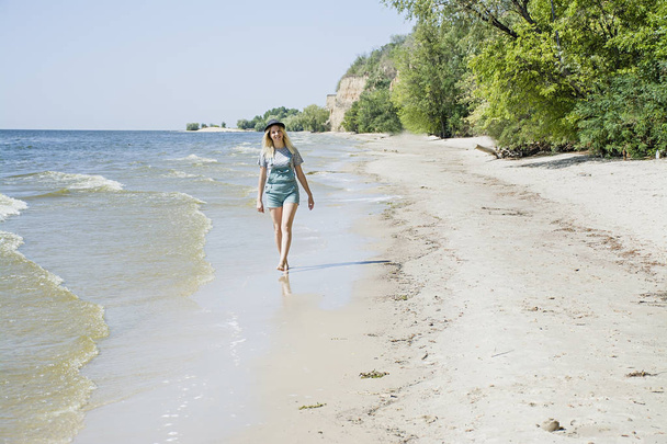 Το κορίτσι στην παραλία φορώντας ένα καπέλο περπατά κατά μήκος της παραλίας, έννοια της ανάπαυσης. Θάλασσα, παραλία, καλοκαίρι. Ώρα χαλάρωσης. - Φωτογραφία, εικόνα