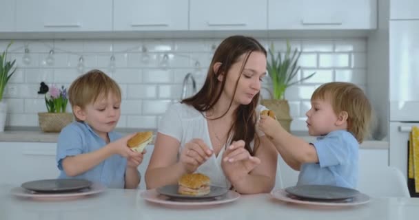 Una joven y hermosa madre en un vestido blanco con dos hijos sonríe y come hamburguesas frescas en su cocina. Familia feliz comida casera alimentos saludables
 - Metraje, vídeo
