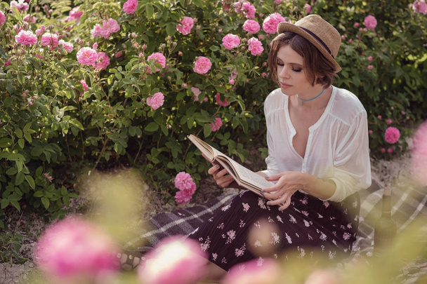 mooi meisje draagt hoed met boek zittend op gras in Rose gaden - Foto, afbeelding