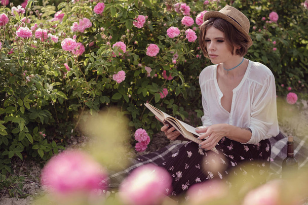 beautiful girl wearing hat with book sitting on grass in rose gaden - Foto, afbeelding