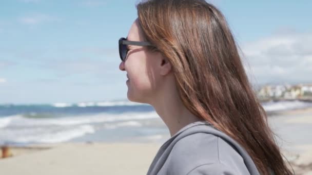 Μια νεαρή γυναίκα κοιτάζει τον ωκεανό. Κοντινό πλάνο του προσώπου της στα γυαλιά ηλίου - Πλάνα, βίντεο