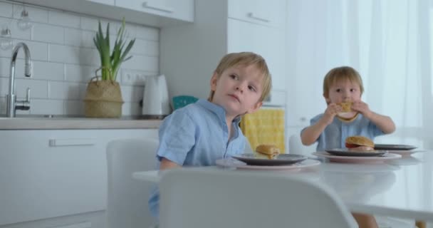 Młoda piękna matka w białej sukni z dwójką dzieci uśmiecha się i zjada świeże hamburgery w kuchni. Szczęśliwa rodzina domowe jedzenie zdrowej żywności - Materiał filmowy, wideo