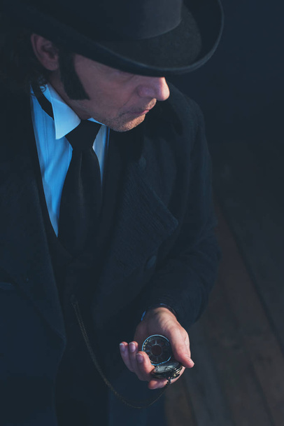Εδουαρδιανός άνθρωπος με μακρύ μαύρο παλτό και καπέλο κρατώντας ρολόι τσέπης. - Φωτογραφία, εικόνα