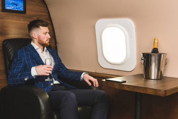 A young successful businessman in an expensive suit sits in the chair of a private jet with a glass of champagne in his hand and looks out the window - 写真・画像