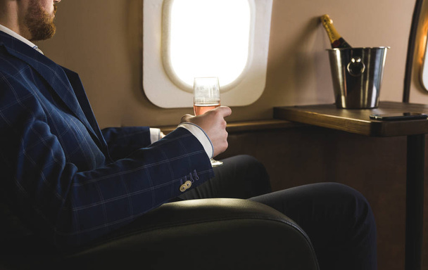 Ένας νέος επιτυχημένος επιχειρηματίας με ακριβό κουστούμι κάθεται στην καρέκλα ενός ιδιωτικού τζετ με ένα ποτήρι σαμπάνιας στο χέρι του και κοιτάζει έξω από το παράθυρο - Φωτογραφία, εικόνα
