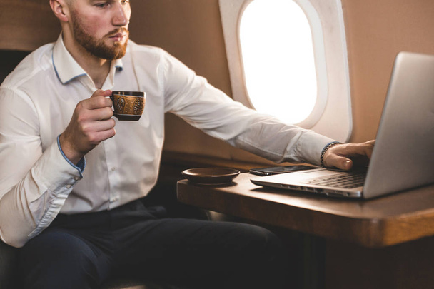 Attractive and successful businessman drinking coffee and working behind a laptop while sitting in a chair of his private jet. - Фото, изображение