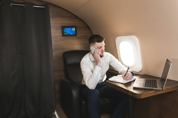 Young attractive and successful businessman talking on the phone and working while sitting in the chair of his private business plane - Foto, Imagen