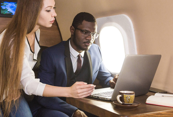 Молодой успешный афро-американский бизнесмен в очках и привлекательная блондинка-коллега в частном самолете. Сервис и перелет первым классом
 - Фото, изображение
